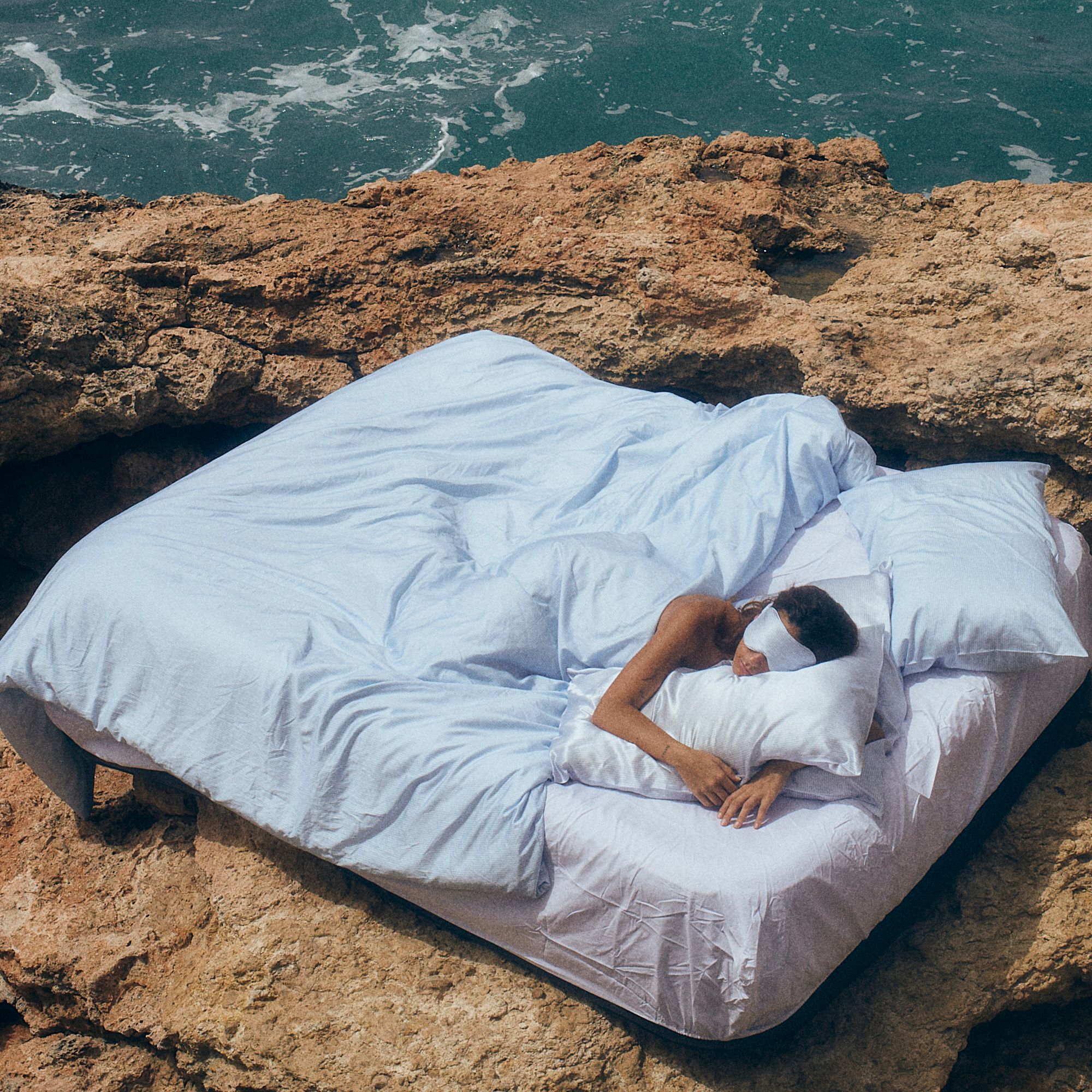 Person lying on a bed with white sheets and pillows on a rocky cliff overlooking the ocean.