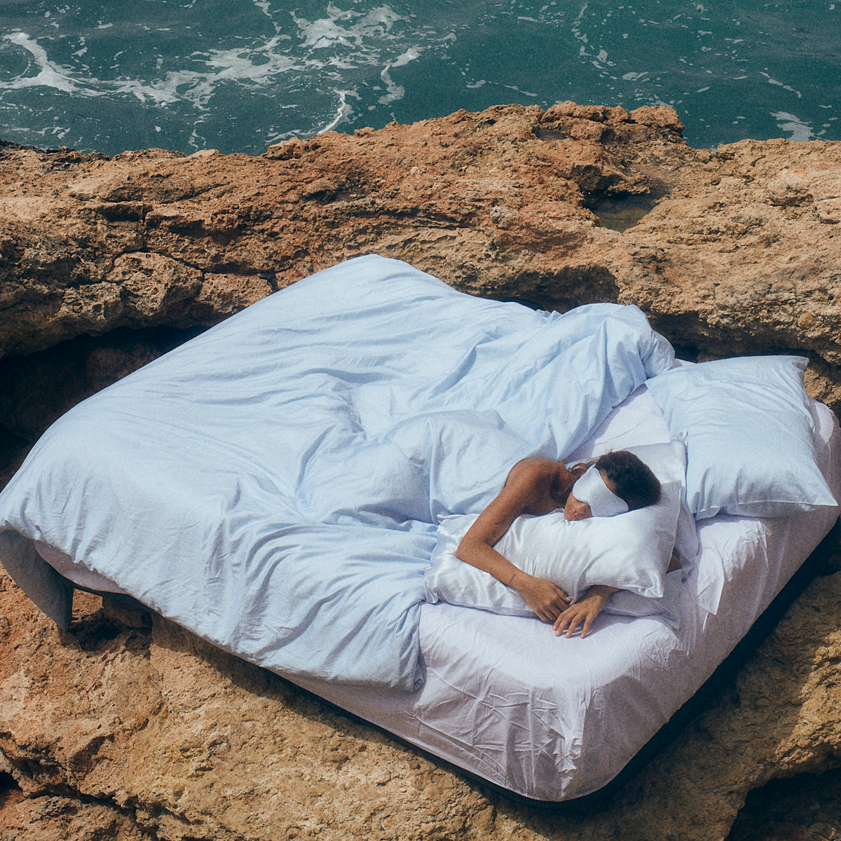 Person lying on a bed with white sheets and pillows on a rocky cliff overlooking the ocean.